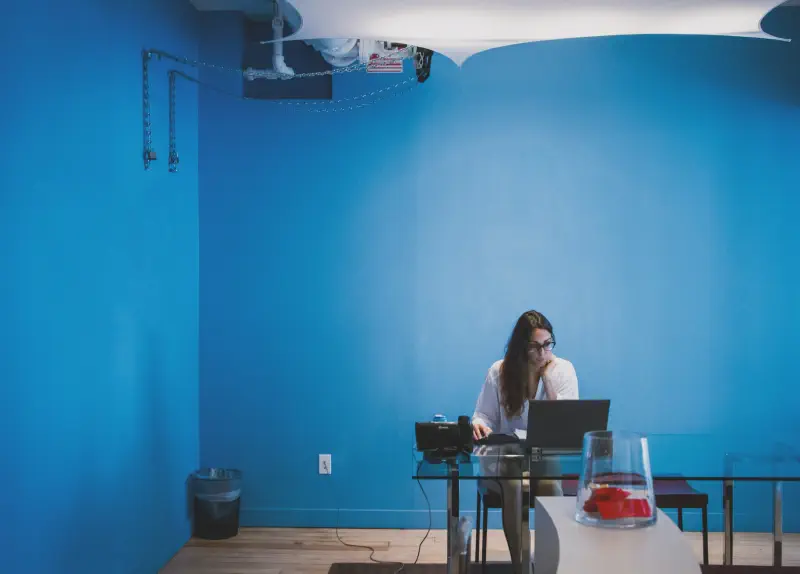 A stock photo of a woman working at a desk, with a bold blue wall behind her.